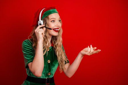 Young female wearing festive green elf costume with headset on vibrant red background, depicting...