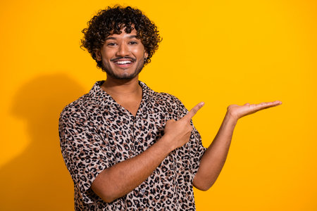 Cheerful young man in leopard print shirt pointing on yellow background ideal for advertising or promotional conceptsの写真素材