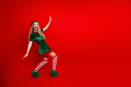 Stylish young woman in festive elf costume dancing joyfully against a vibrant red backdrop during Christmas timeの写真素材