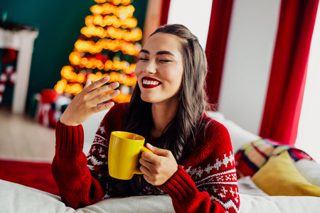 Woman Enjoying a Cozy Christmas Morning with a Bright Yellow Cup and a Beautifully Lit Festive Tree at Homeの写真素材