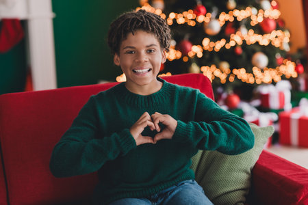 Happy young boy wearing a green sweater making a heart sign while seated indoors near a beautifully decorated Christmas treeの写真素材