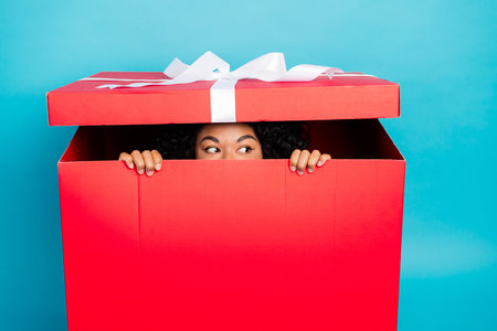 Cheerful woman peeking out of a giant bright red gift box against a blue background, evoking surprise and holiday spiritの写真素材