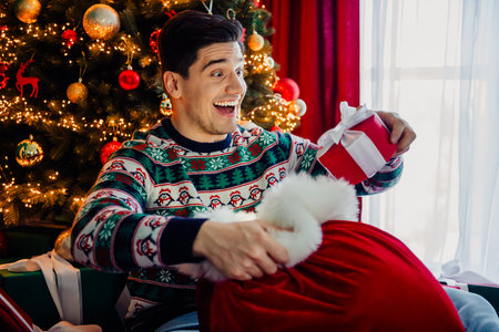 Joyful young man unwrapping Christmas gift near a festive tree, capturing the charm of the holiday seasonの写真素材