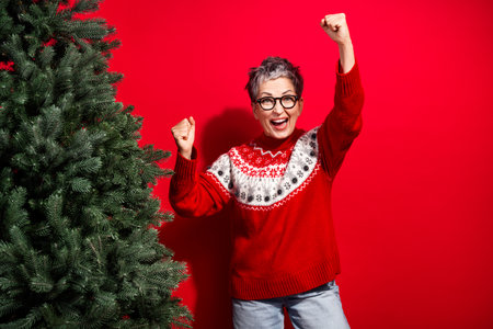 Joyful mature woman in festive red sweater celebrating Christmas near a tree against a bright red backgroundの写真素材