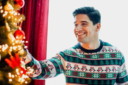 Smiling young man in festive sweater decorating Christmas tree with ornaments in cozy holiday atmosphereの写真素材