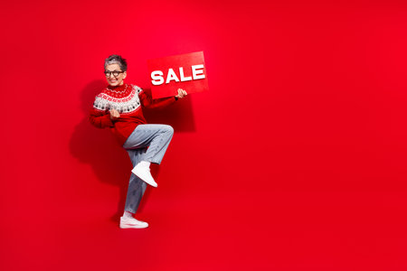 Mature woman in festive sweater jumps joyfully while holding a sale sign against a vibrant red backgroundの写真素材