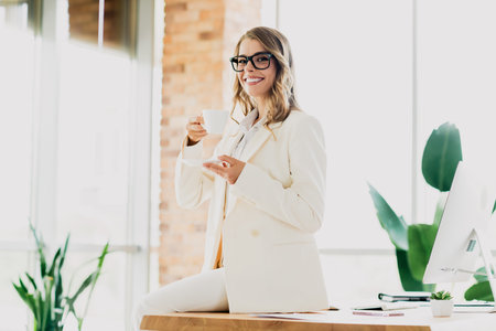 Smiling professional businesswoman in formal attire enjoying a coffee break in a bright modern office settingの写真素材