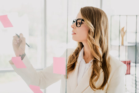Focused businesswoman in formal attire writing ideas on sticky notes in a modern office environmentの写真素材