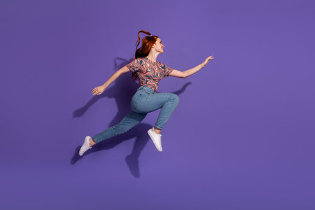 Energetic young red-haired woman leaping joyfully against a solid purple background, showcasing vibrant casual fashion and styleの写真素材