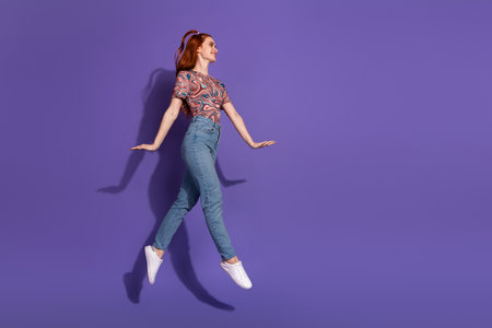 Young red-haired woman jumping on a vibrant violet background wearing casual clothes exuding positivity and energyの写真素材