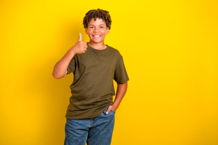 Cheerful young boy in casual khaki tshirt and jeans gives thumbs up against a bright yellow background ideal for lifestyle and advertisingの写真素材