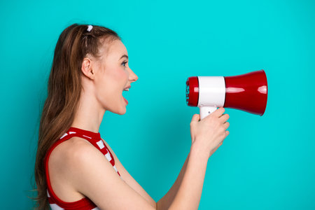 Positive woman using a red megaphone while expressing excitement and joy against a vibrant teal backgroundの写真素材