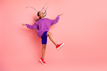 Young joyful woman in a purple hoodie and shorts jumping energetically against a pink background, showcasing vibrant styleの写真素材