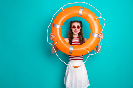 Young woman holding an orange lifebuoy against a vibrant teal background, wearing stylish striped clothingの写真素材