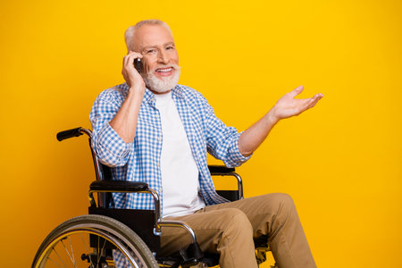 Grandpa in a wheelchair on a bright yellow studio backdrop talks on his phone with a warm smile and open hand inviting inclusion accessibility and care in everyday lifeの写真素材
