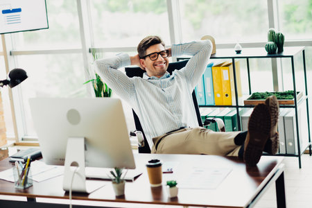 Businessman relaxing confidently in a modern office workspace surrounded by technology and stylish decor on a productive dayの写真素材