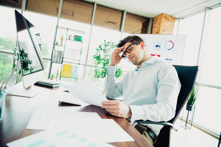 Professional businessman analyzing paperwork at modern office desk with focus and determination during a work dayの写真素材