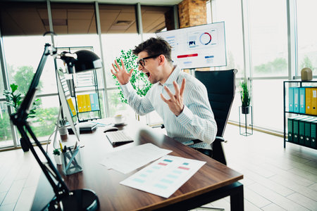 Young businessman expressing frustration at work in a modern office setup, surrounded by financial documents and reportsの写真素材