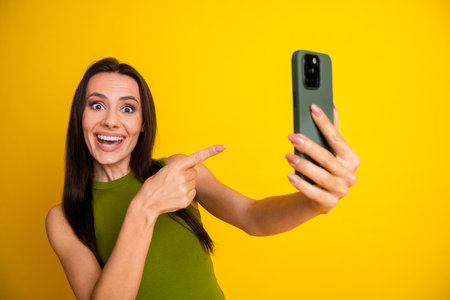 Smiling woman holding a smartphone against a bright yellow background, wearing a green top and exuding happinessの写真素材