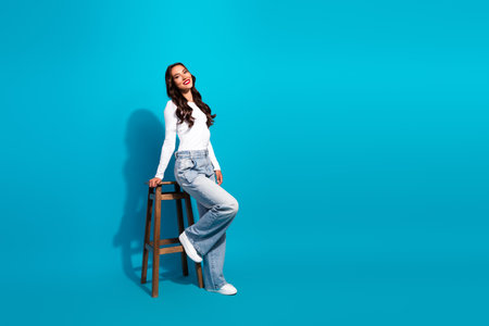 Smiling young woman posing stylishly in casual fashion with a wooden stool against a vibrant blue backgroundの写真素材