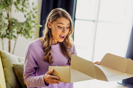 Young woman opening a package in a cozy living room, showing surprise and joyの写真素材