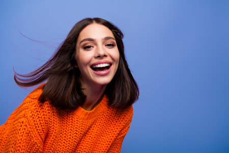 Smiling young woman in an orange sweater with vibrant purple background showcasing happiness and modern fashion styleの写真素材