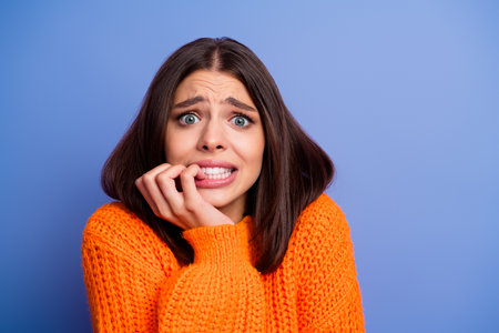 Young woman in bright orange sweater expressing anxiety with a concerned look against a plain blue-purple background.の写真素材