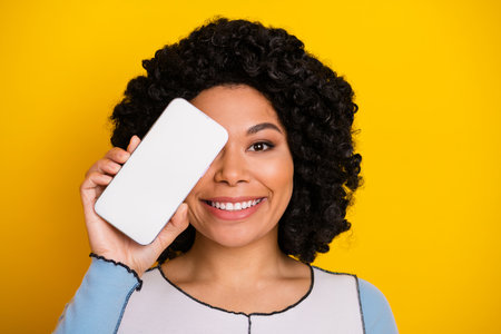 Smiling young woman holding a smartphone over bright yellow background, promoting technology and digital communicationの写真素材