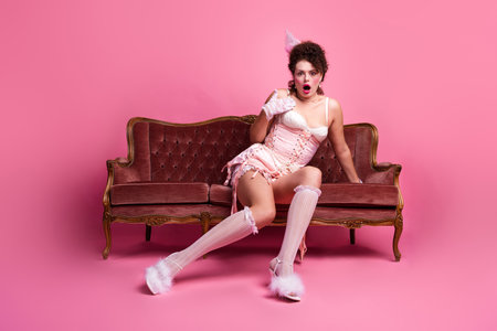 A young woman dressed in a vintage-inspired corset and socks sits on an antique sofa with a surprised expression against a pink backgroundの写真素材