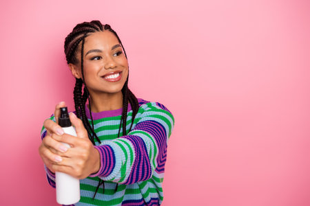 Young woman with radiant smile spraying from bottle, posing in colorful sweater on pink background, promoting beautyの写真素材