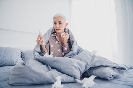 Charming elderly woman with white hair sitting in bed holding tissues, wearing pajamas, reflecting natural daylightの写真素材