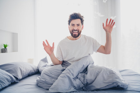 Photo of cheerful crazy young man dressed nightwear sititng bed having fun indoors house apartment roomの写真素材