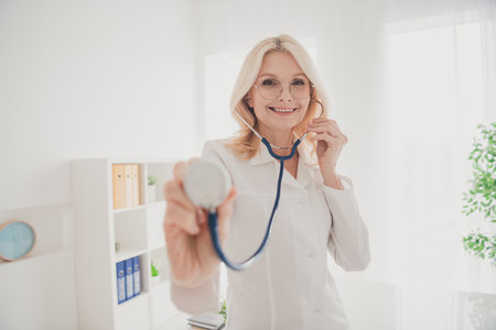 Senior female doctor in a clinic office smiles while holding a stethoscope, reflecting healthcare professionalismの写真素材