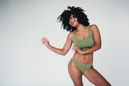 Young woman with curly hair posing confidently in stylish lingerie against a neutral studio backgroundの写真素材