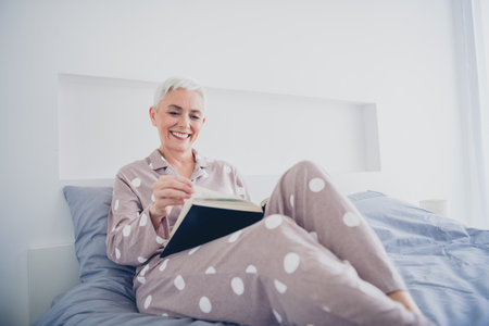 Elderly woman in pajamas enjoying reading in bed, capturing the essence of leisure and comfort in a bright, tranquil bedroom settingの写真素材