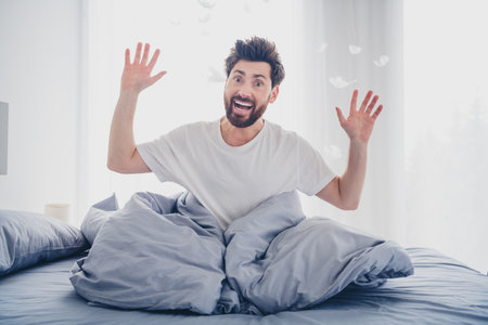 Photo of impressed funny young man nightwear pajama sitting bed rising arms indoors house apartment roomの写真素材