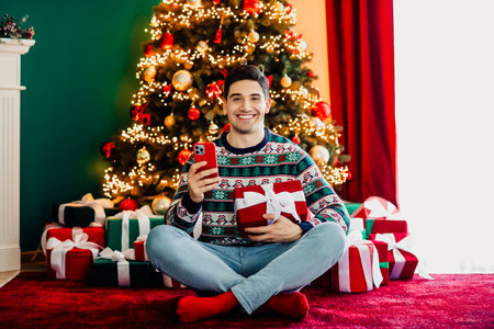 Cheerful man holding a smartphone and a gift in a festive setting with a decorated Christmas tree and glowing lightsの写真素材