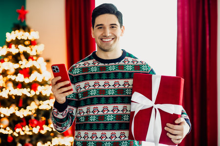 Young man celebrating Christmas holding a smartphone and present in a cozy holiday setting with...