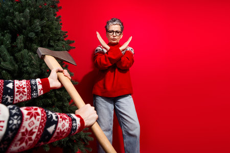Senior lady in red sweater showing cross hand gesture rejecting Christmas tree chopping idea on bright festive backgroundの写真素材