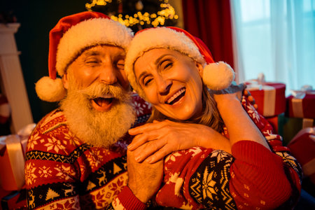 Joyful Christmas couple in festive sweaters laughing together by a warm tree with gifts at home in a cozy holiday sceneの写真素材