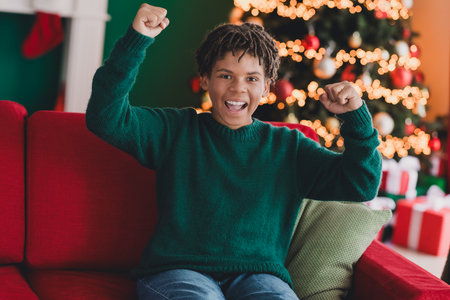 Excited boy in green sweater celebrating Christmas, sitting on a red sofa near a lit tree in a cozy festive room.の写真素材