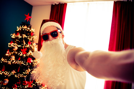 Festive selfie of a person dressed as Santa Claus in modern style near a beautifully lit Christmas tree showcasing holiday cheerの写真素材
