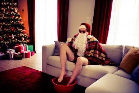 Santa Claus relaxing in a festive living room with presents, a lit Christmas tree, and cozy winter decorationsの写真素材