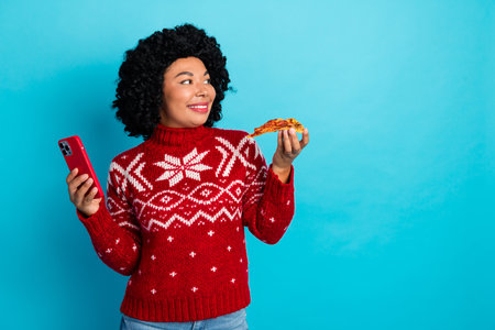 Smiling woman in festive red sweater holding pizza slice and smartphone against blue background celebrating holiday momentの写真素材