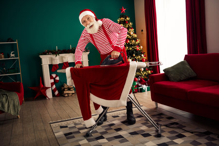 Santa Claus preparing his iconic red suit by ironing during Christmas time in a festive indoor setting with holiday decorの写真素材
