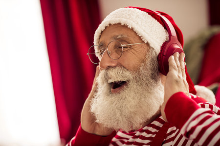 Elderly Santa Claus Enjoying Christmas Music Indoors Wearing Headphones and Red Hat in a Festive Winter Settingの写真素材