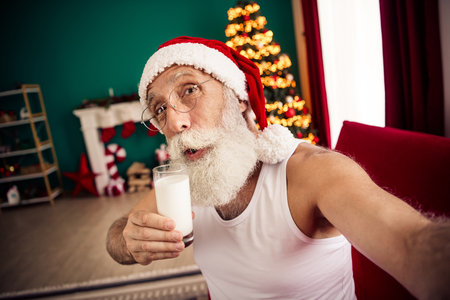 Festive senior man dressed as Santa Claus enjoying milk at home near a decorated Christmas treeの写真素材