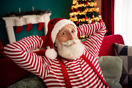 Cheerful man dressed as Santa Claus sitting by a festive Christmas tree and fireplace in a colorful and cozy homeの写真素材
