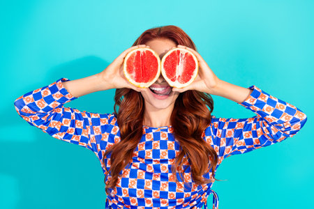 Cheerful young woman holding fruit halves playfully over her eyes, styled in a trendy outfit against...