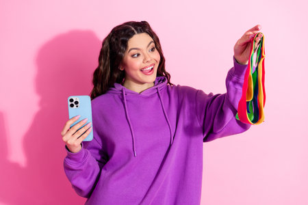 Pretty woman in purple sweatshirt holding colorful fitness bands and smartphone on a pink background demonstrating excitementの写真素材